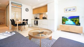 A modern living room with a sofa, coffee table, and TV. There's a dining area with chairs and a table, and a kitchen with cabinets and appliances. - Resort-Style 1BR Apt in Adelaide CBD | Pool & Spa (Adelaide)