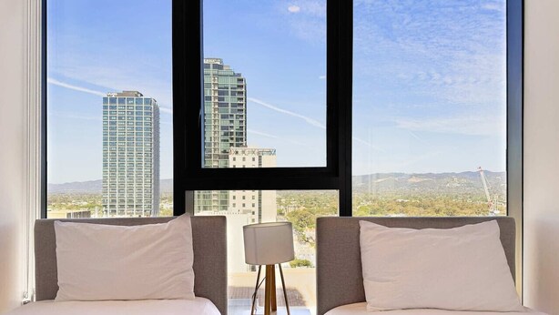 Habitación de hotel con dos camas, una mesita de noche y una lámpara. La habitación tiene ventanas amplias que ofrecen vistas a edificios de la ciudad y un paisaje distante.