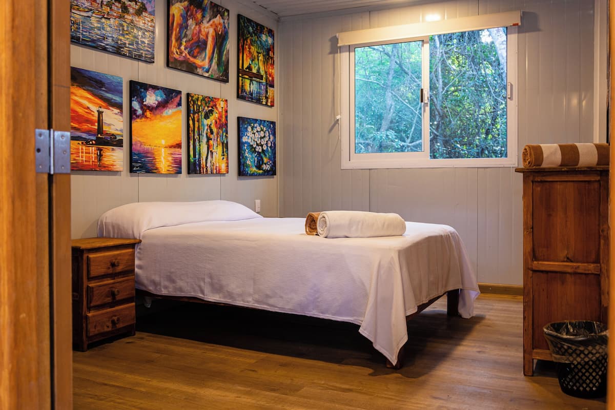 A bedroom with a bed, wooden dresser, and a window with a view of trees.