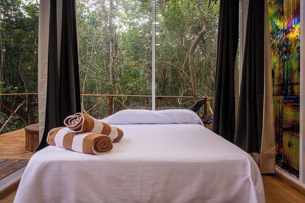A bed with white linens and brown striped towels, a wooden deck, and a view of a forest through large windows.
