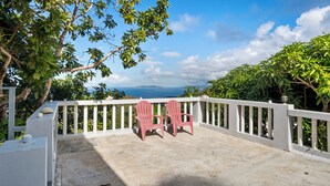 Ein Balkon mit zwei roten Adirondack-Stühlen, einem weißen Geländer und Blick auf das Meer und die Berge.