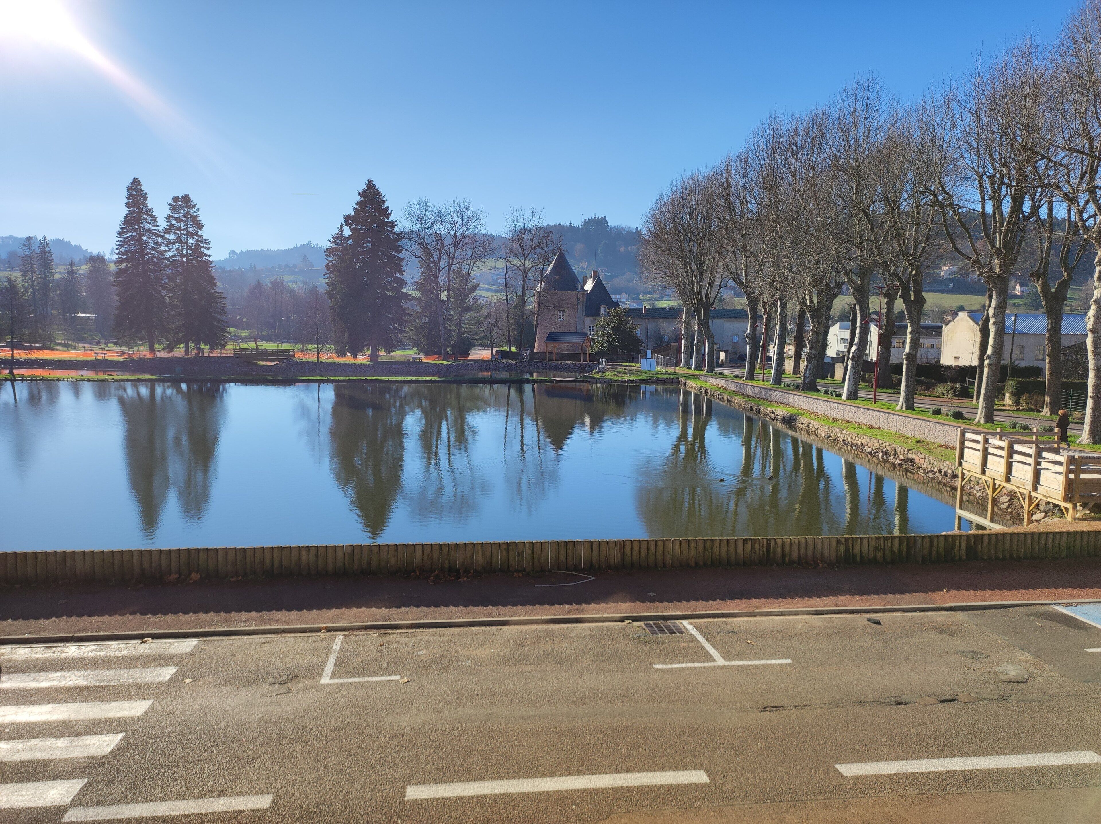Un lac paisible dont les eaux reflètent les arbres et un bâtiment situé au loin, le tout entouré d’un parc et d’une route sur laquelle se trouve un pont piétonnier.