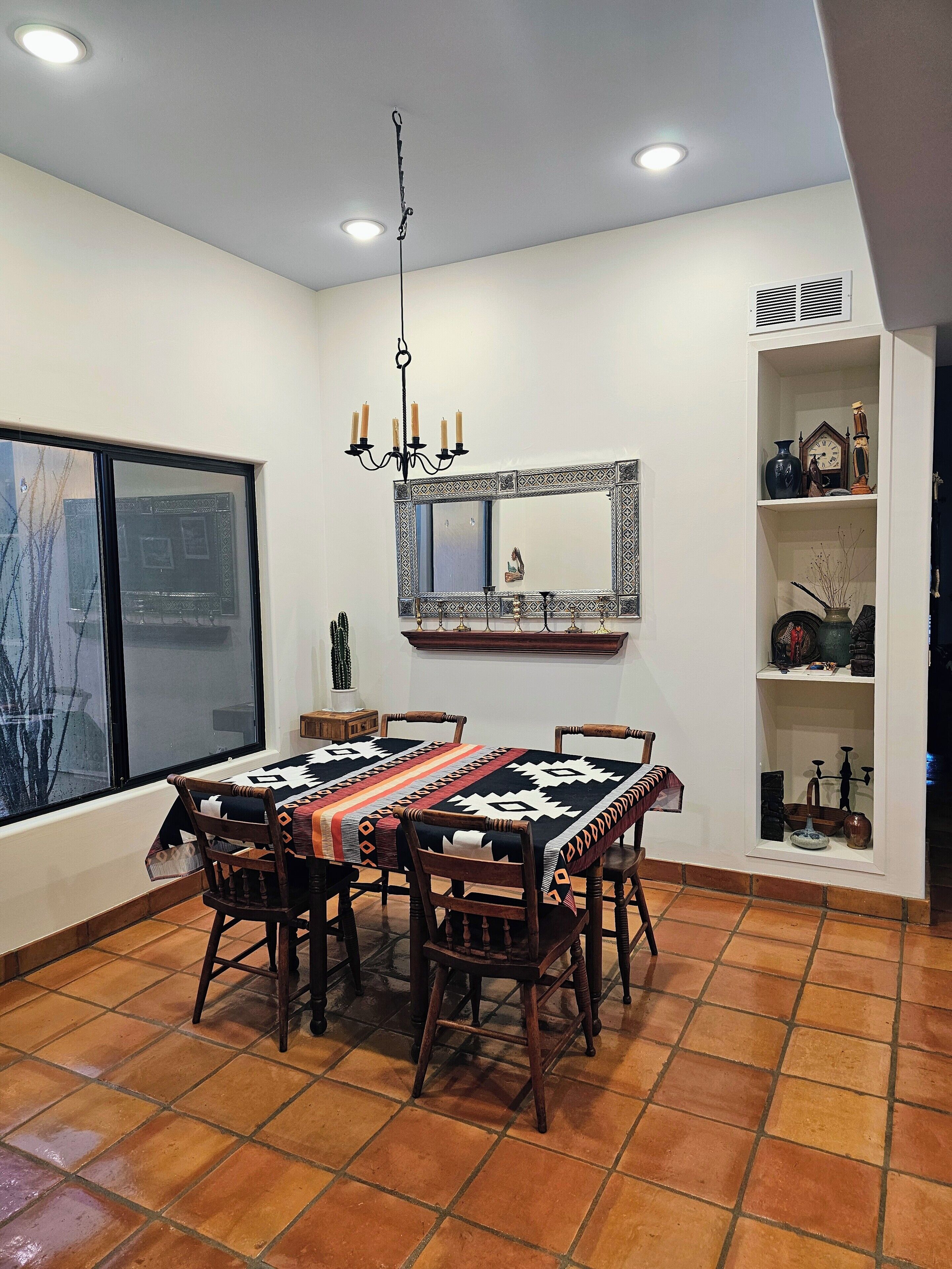 A dining area with a large table and chairs, a chandelier, a mirror with decorative frame, and a shelf with various items.