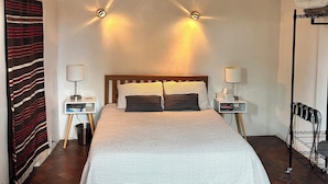 A bedroom with a bed, two bedside tables with lamps, a wooden headboard, a striped curtain, a small table with a lamp, and a chair.