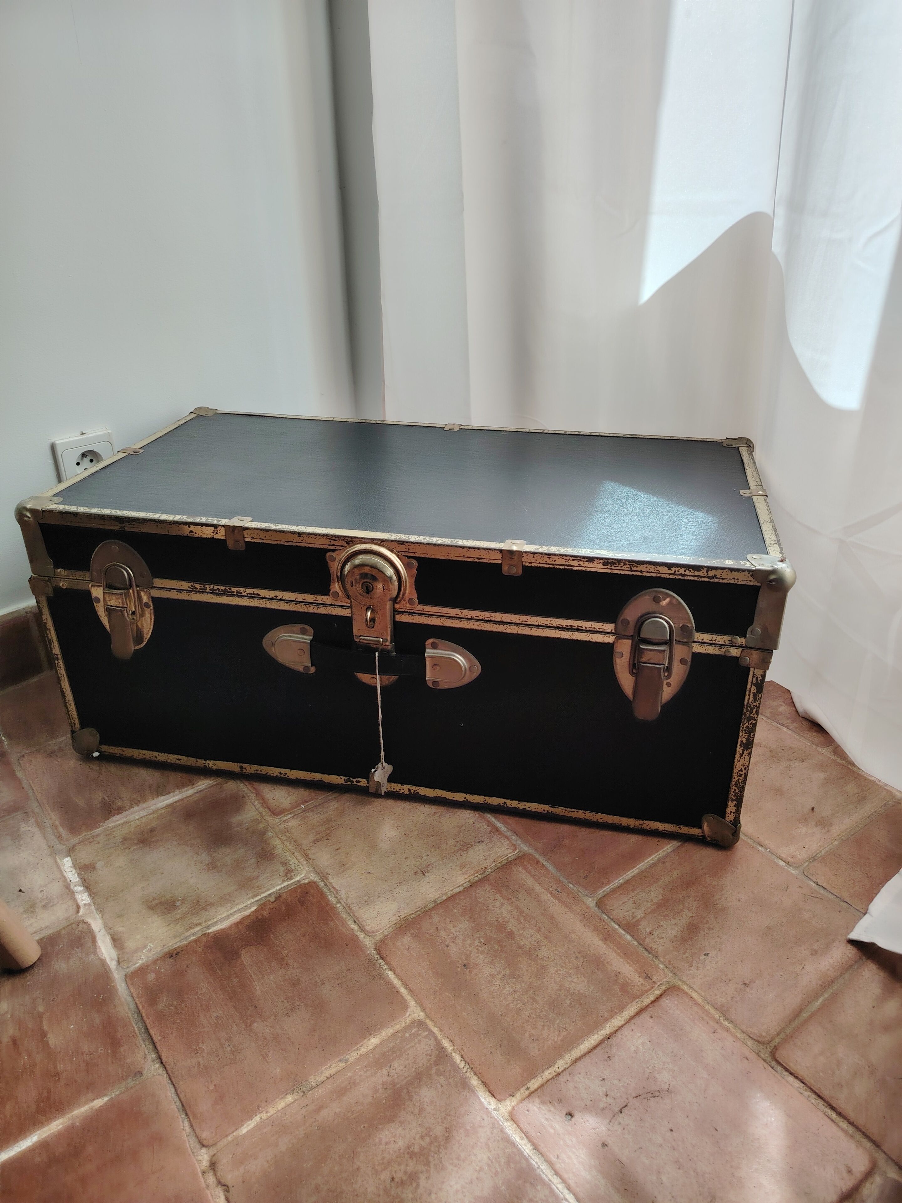 Een zwarte vintage koffer met metalen sluitingen en een handgreep, op een betegelde vloer voor een witte muur.