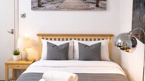 A neatly made bed with a wooden headboard, a bedside lamp, and a rolled towel on the bed. - Paddington Studios • by Oxford Street & Marylebone (London)