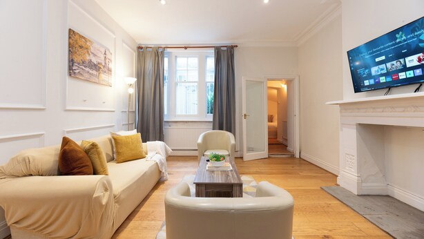 A modern living room with a sofa, armchair, and a flat-screen TV mounted above a fireplace. - Two Bedroom Flat - Next to West Kensington Station - Pass the Keys (London)