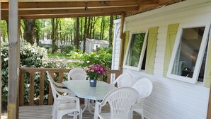 Un patio exterior techado con piso de madera, sillas de plástico blanco y una mesa con planta en maceta.