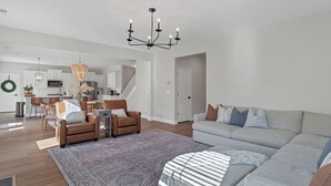A spacious living room with a large gray sectional sofa, a brown leather chair, and a patterned rug. A kitchen with white cabinets and a hanging light fixture is visible in the background. - Spacious 6-bedroom house near Pinehurst (Southern Pines)