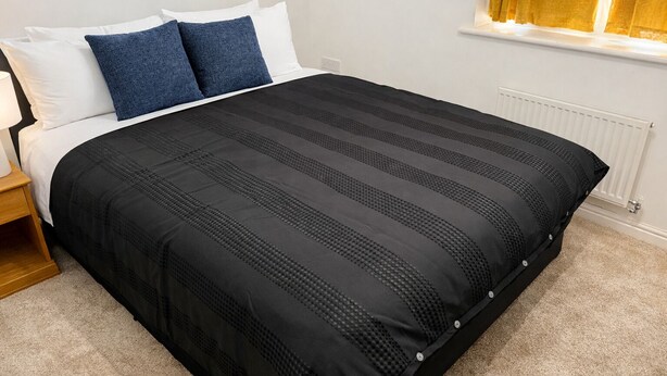 Een net opgemaakt bed met een zwart-wit gestreept dekbed, een houten nachtkastje en een raam met gele gordijnen.