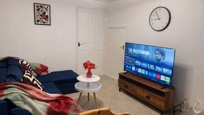 Een woonkamer met een flatscreen televisie waarop 'The Mastermind' te zien is, een blauwe bank, een klein rond tafeltje met een vaas en een houten stoel.
