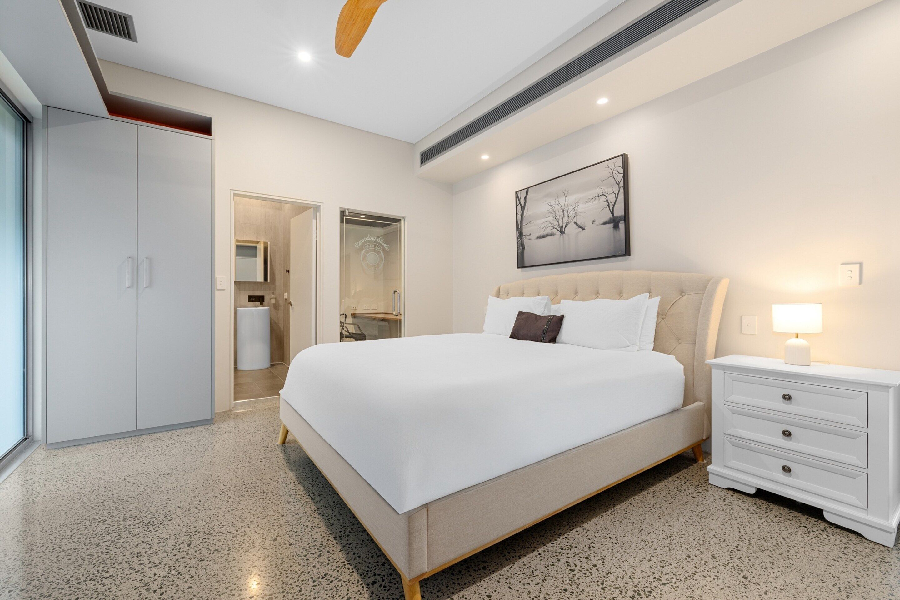 Un dormitorio moderno con una cama grande, una mesita de noche, un armario y un baño visible a través de una puerta abierta.