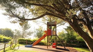 A playground with a slide and climbing structure, surrounded by trees and grass. - Mobilheim auf dem Hugel mit Pool (Villanova d'Albenga)