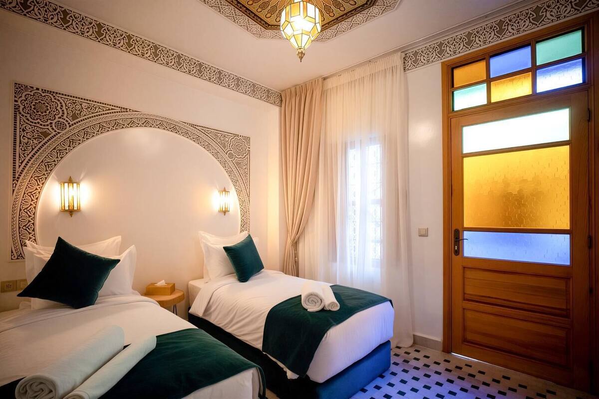 A hotel room with two beds, a chandelier, a wooden door with frosted glass panels, and patterned wall decor.
