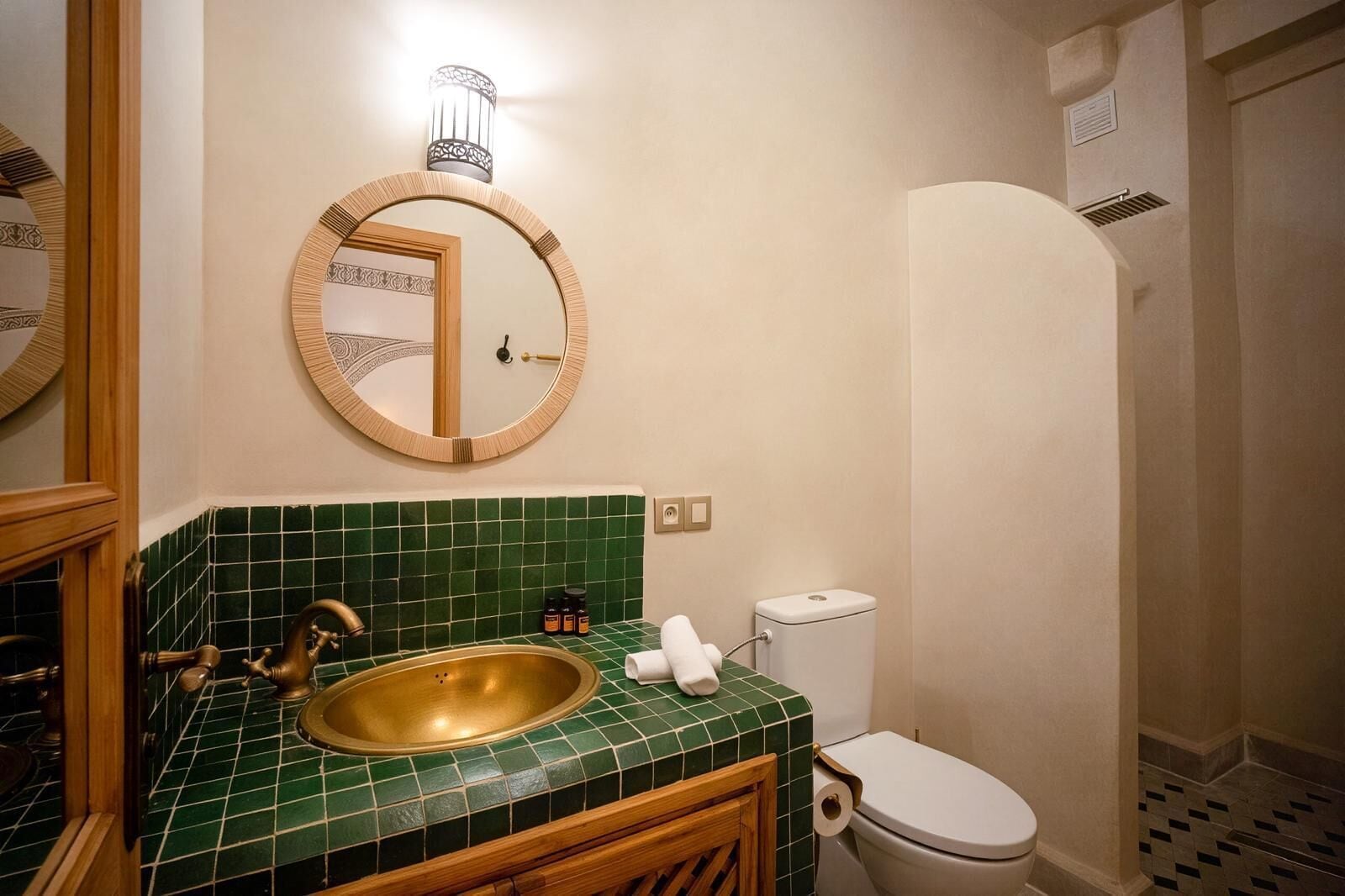 Un baño con lavamanos con azulejos verdes, grifo de latón, inodoro blanco y zona de ducha.