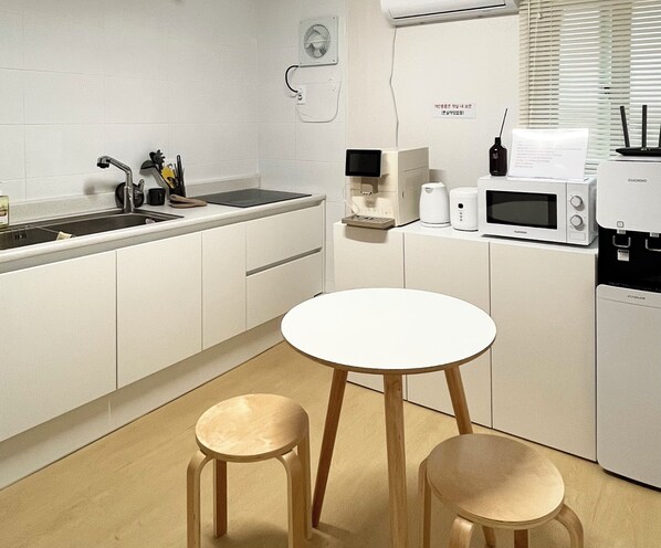A modern kitchen with white cabinetry, a round white table, two wooden stools, a built-in sink, and various appliances including a microwave and coffee maker.
