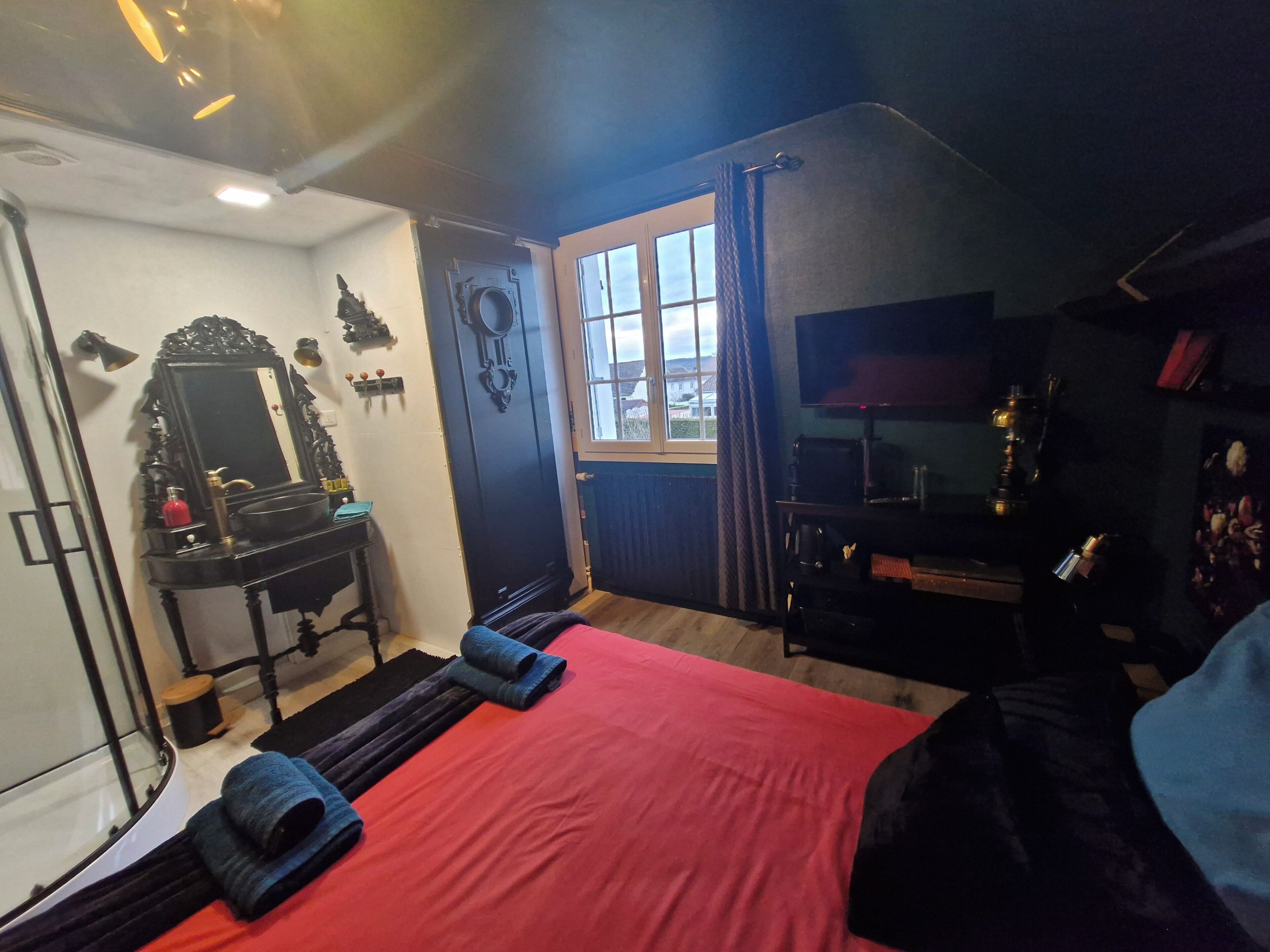 Une chambre d’hôtel avec un lit rouge, un bureau noir avec un miroir, une télévision et une fenêtre avec des rideaux.