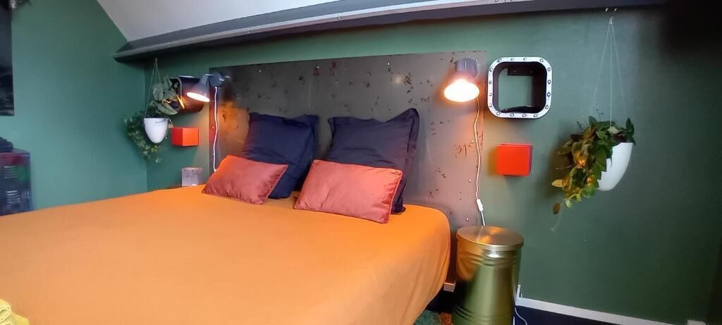 Sänky, jossa on oranssit vuodevaatteet, kaksi tyynyä, sisäänrakennetulla valaistuksella varustettu pääty ja kaksi seinälle kiinnitettyä kasvia.