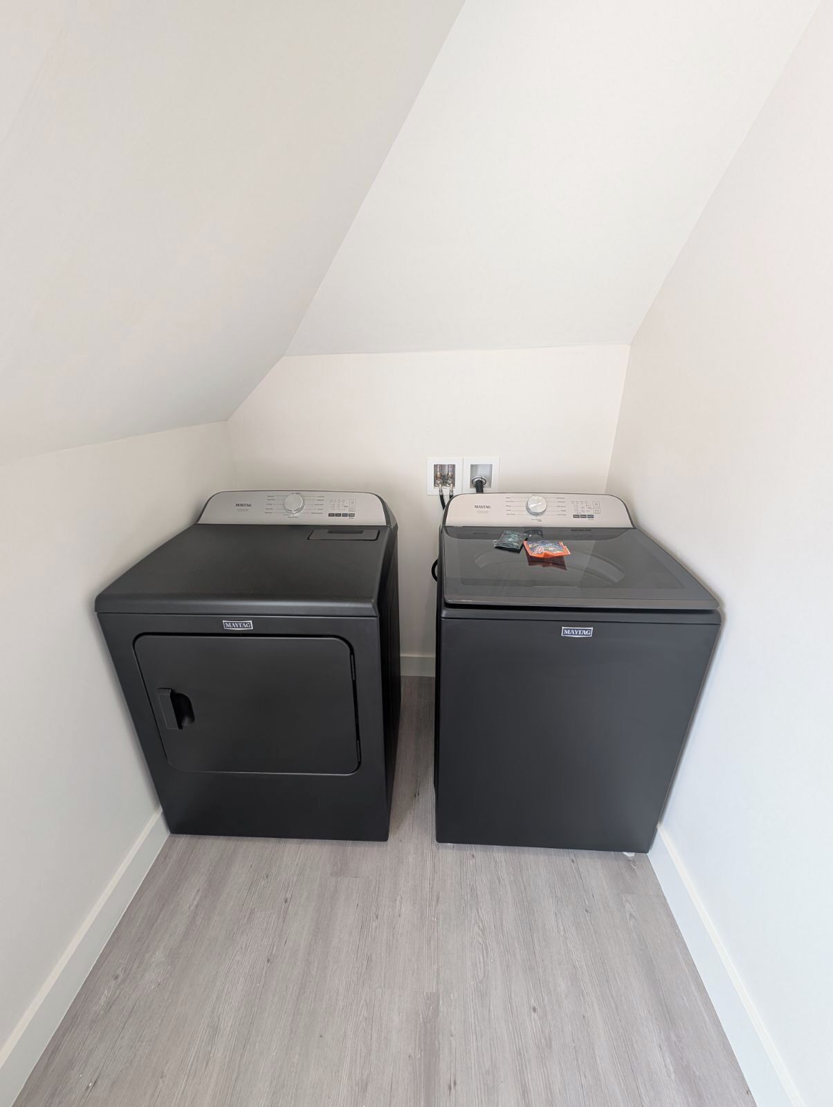 Deux machines à laver Maytag noires, placées dans un coin d’une pièce aux murs blancs et au plafond en pente.