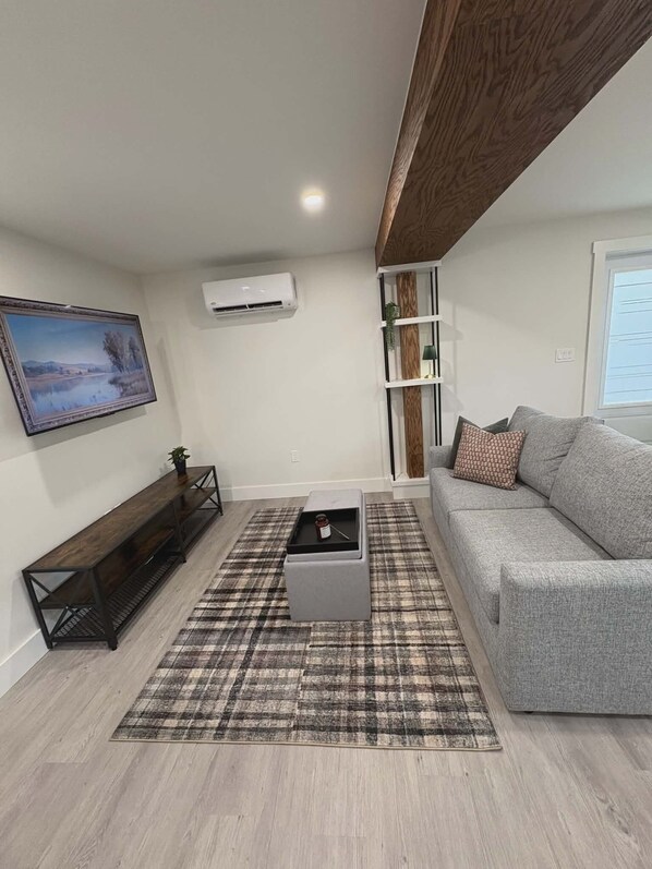 Una sala moderna con un sofá gris, un banco de madera, un tapete escocés y una pequeña mesa con un tarro.