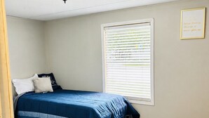 Un dormitorio con cama, ventana con persianas, luz de techo y una frase motivacional en la pared.