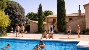 Ein Schwimmbecken mit Personen darin und darum herum, umgeben von Steinbauten und Bäumen.