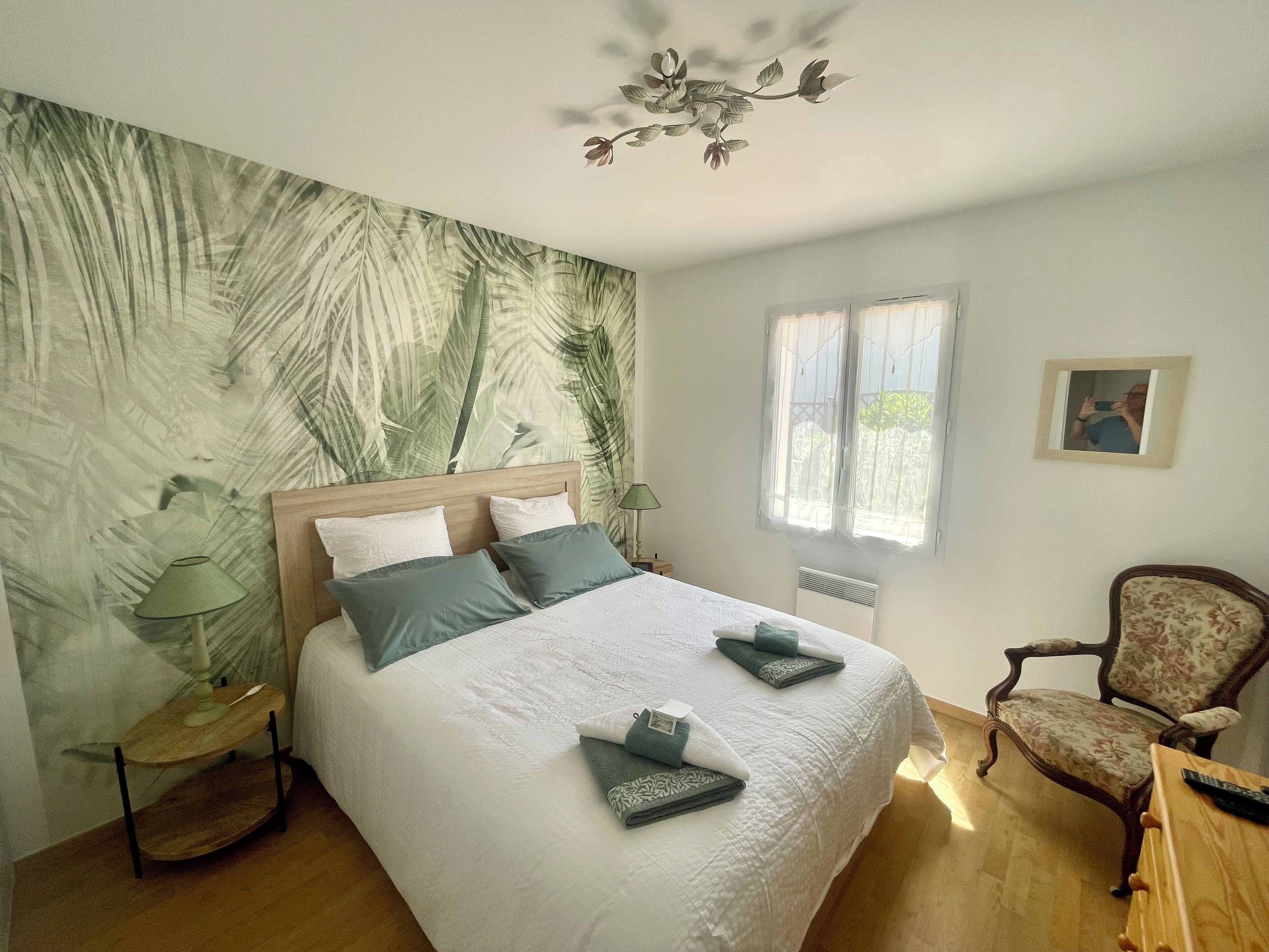 Un dormitorio con cama, una silla, una lámpara, un ventanal y una pared con diseño de hojas.