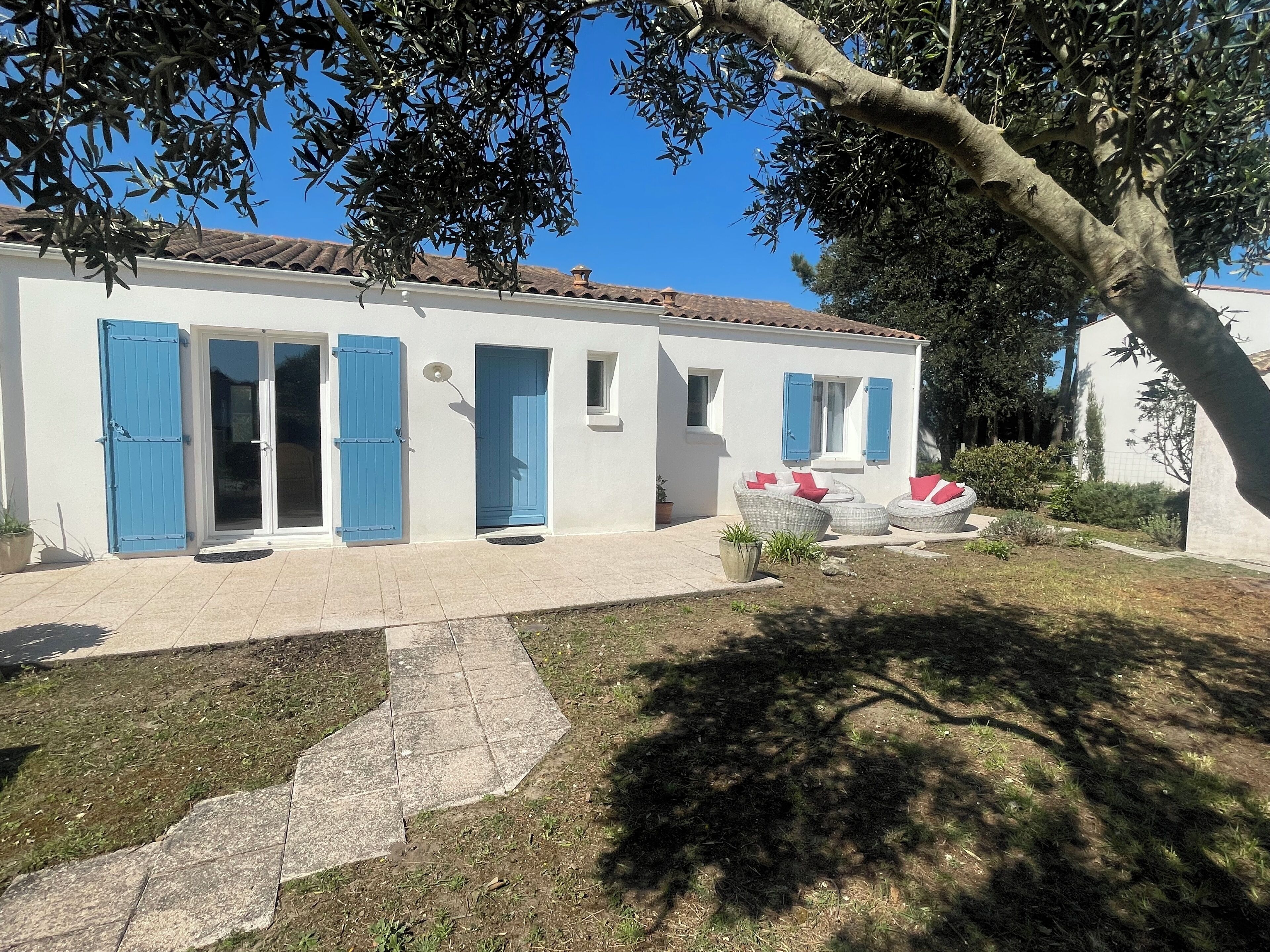 Una casa de dos pisos con contraventanas azules, un patio con baldosas y mobiliario exterior.