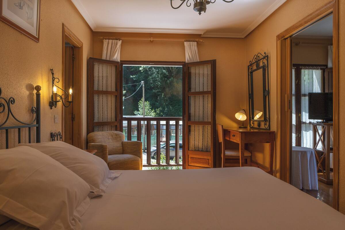 A bedroom with a large bed, a chair, a desk with a lamp, and a mirror. There is a window with curtains and a view of greenery outside.