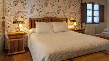 A bedroom with a wooden headboard, two bedside tables with lamps, a large bed with white bedding, floral wallpaper, and a window with wooden shutters.