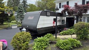 Ein Wohnmobil der Marke Coleman vor einem Haus mit einem Basketballkorb, umgeben von gepflegten Büschen und einem kleinen Garten.