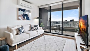 Un salon moderne avec un canapé, un téléviseur et une vue sur le paysage urbain, visible à travers de grandes portes coulissantes.
