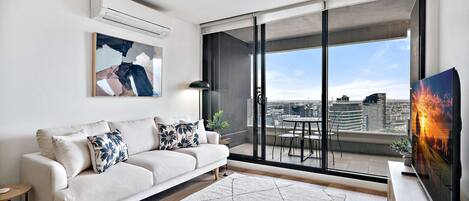 A modern living room with a sofa, a TV, and a view of a cityscape through large sliding doors.