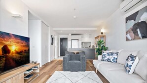 Un salon moderne avec un téléviseur à écran plat, un fauteuil gris, un canapé blanc agrémenté de coussins à motifs floraux, et une cuisine en arrière-plan.