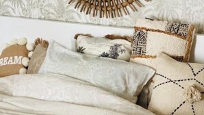 Ein Bett mit einer beigefarbenen Bettdecke und mehreren Kissen, ein Holzbilde über dem Bett und eine gemusterte Tapete im Hintergrund.
