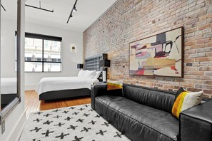 A modern bedroom with a brick wall, a large bed, a black leather sofa, and a city view. - Refined Historic 2BR/1BA Suite in Prime Downtown Wilmington (Wilmington)