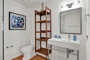 A bathroom with a white toilet, a wooden shelf unit, a white sink with a mirror, and a framed abstract painting on the wall. - Refined Historic 2BR/1BA Suite in Prime Downtown Wilmington (Wilmington)
