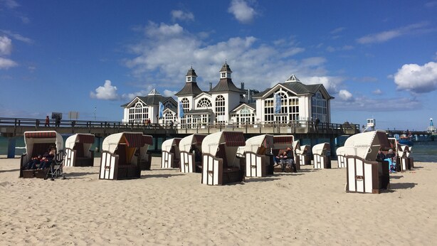 Un edificio frente a la playa con múltiples ventanas y un estilo arquitectónico particular, rodeado de sillas y sombrillas de playa.