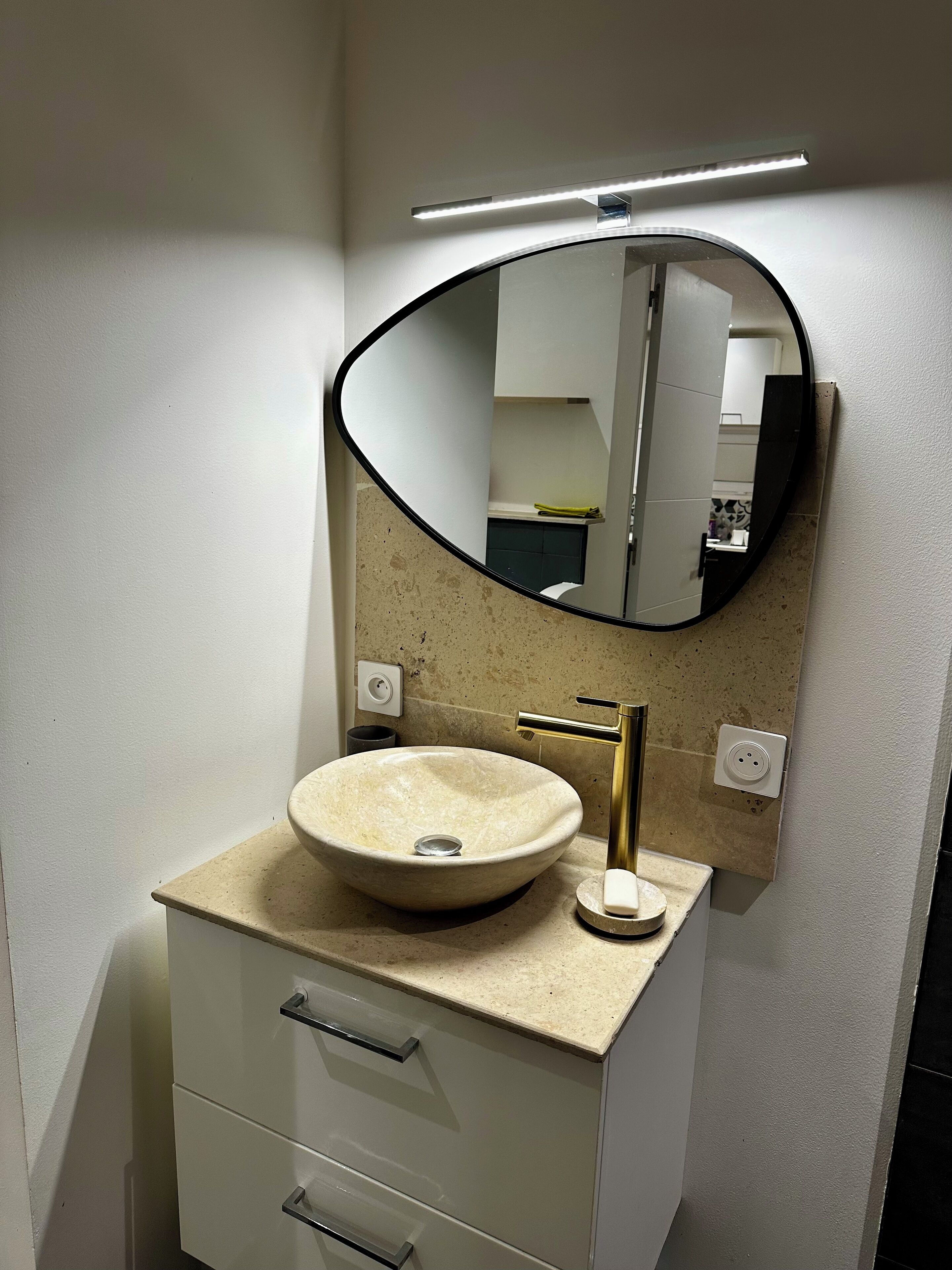 Une salle de bain avec un miroir rond, un lavabo beige, un robinet doré et un meuble blanc avec des tiroirs.