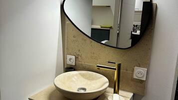 Une salle de bain avec un miroir rond, un lavabo beige, un robinet doré et un meuble blanc avec des tiroirs.