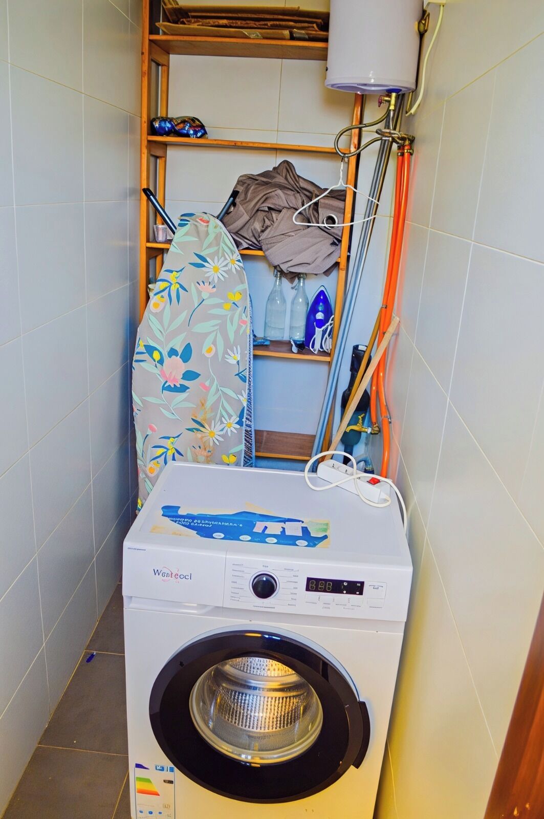 Un lavadero pequeño con una lavadora de carga frontal, un tendedero y un estante con botellas y un objeto azul.