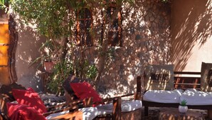 An outdoor seating area with wooden lounge chairs and cushions, a stone wall, and a tree providing shade. - Masairiatacama.cl (San Pedro de Atacama)