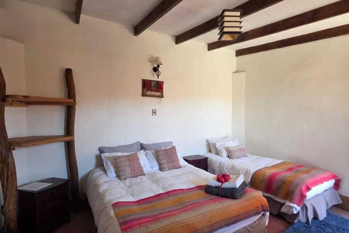 A room with two beds, wooden beams on the ceiling, a small table, and a wall-mounted lamp.