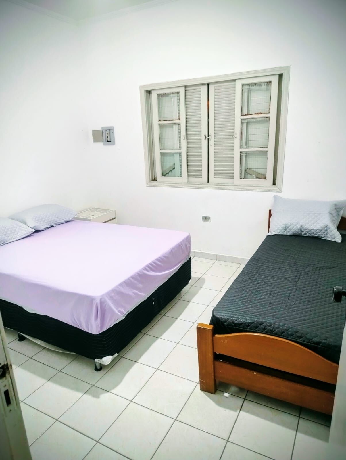 Ein Zimmer mit zwei Betten, eines mit einer violetten Tagesdecke und das andere mit einer schwarzen. Ein Fenster mit weißen Fensterläden und eine kleine Steckdose an der Wand sind ebenfalls vorhanden.