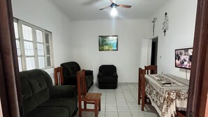 A living room with a green sofa, a wooden chair, a black armchair, a dining table with a tablecloth, a ceiling fan, and a framed picture on the wall. - Complete House with Private Pool and BBQ | Praia Grande (Praia Grande)