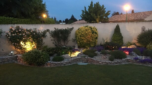 Un giardino curato con un muro in pietra, diversi arbusti e un vialetto ben illuminato.