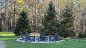 Et cirkulært udendørs opholdsområde med Adirondack-stole og et bålsted, omgivet af en græsplæne og høje træer.