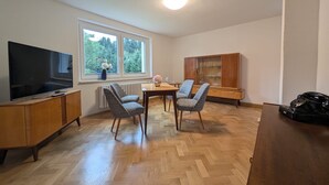 Un salon moderne doté d’un téléviseur à écran plat, d’une table à manger avec des chaises, d’un meuble en bois et d’une grande fenêtre offrant une vue sur les arbres.