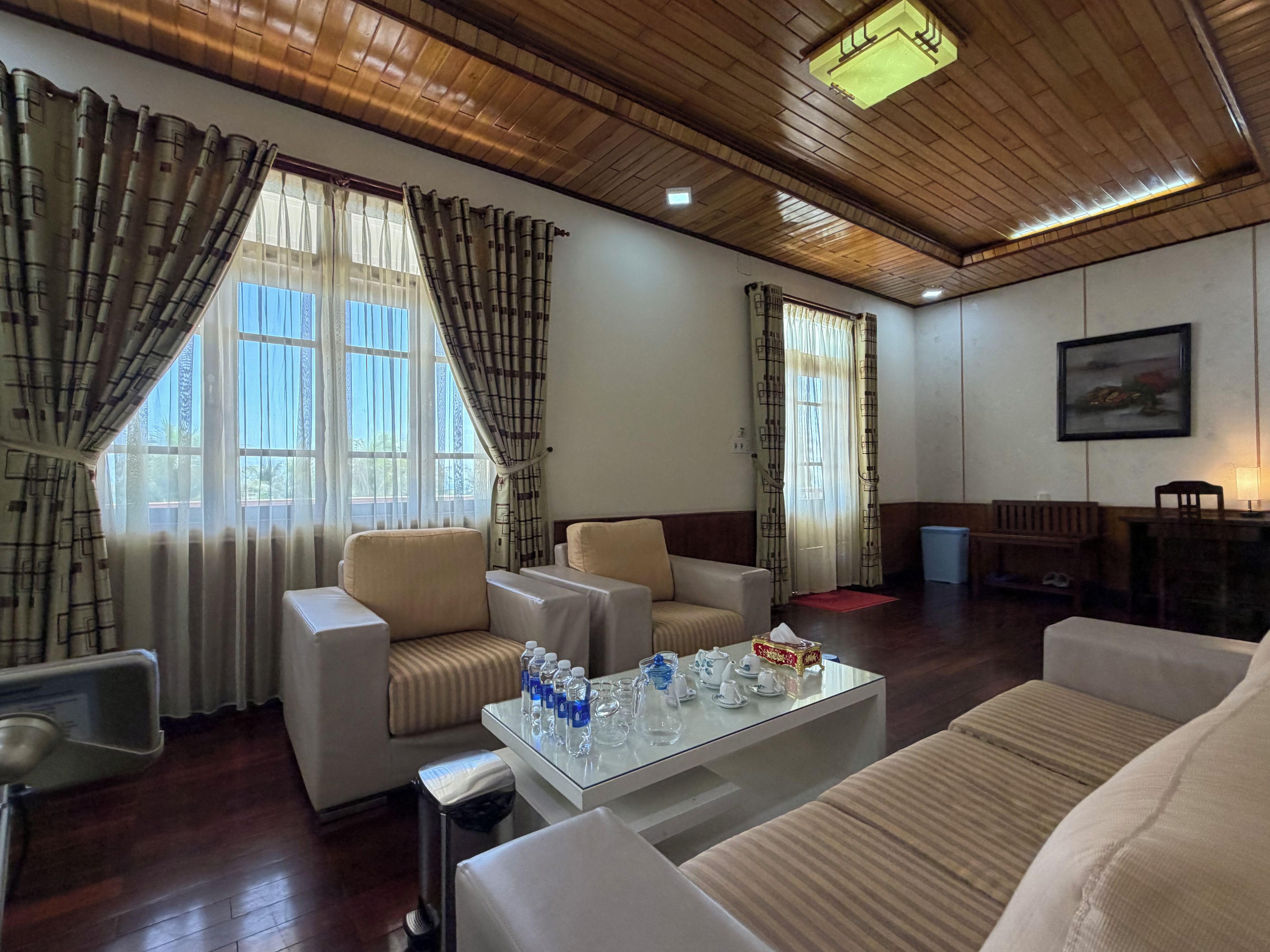 A hotel room with a wooden ceiling, two windows with curtains, a sofa, a table with water bottles, and a painting on the wall.