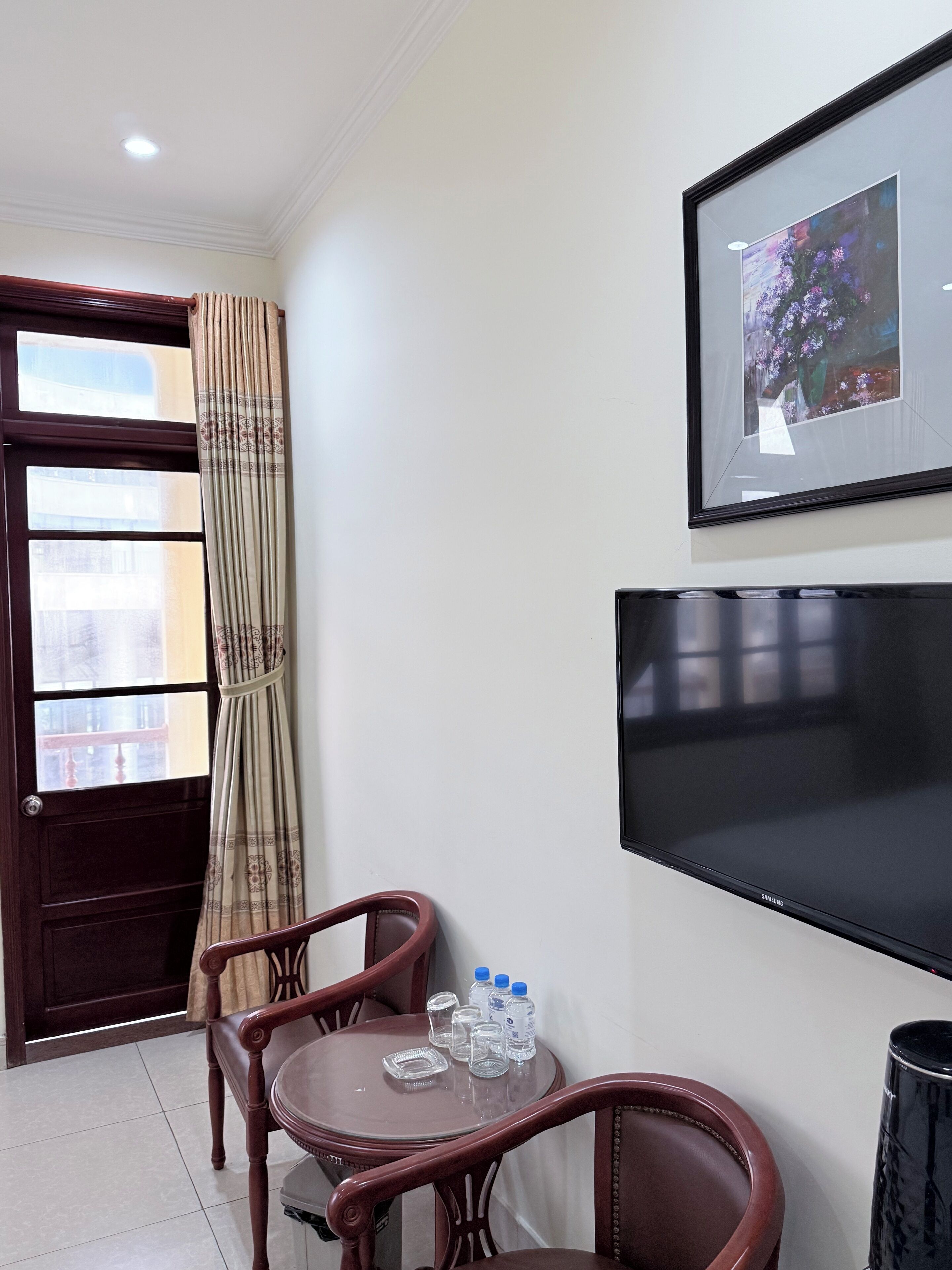 Habitación de hotel con puerta de madera, cortina, cuadro en la pared, televisor de pantalla plana, pequeña mesa con sillas y un cesto de basura.